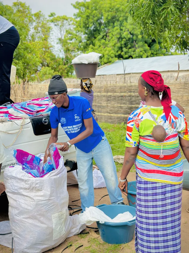 Omar in a blue campaign shirt unpacking a sack of mosquito nets beside a pickup, a woman with a baby on her back in front