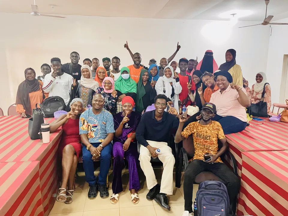 A group photo of about thirty volunteers in a training hall, many holding up one finger