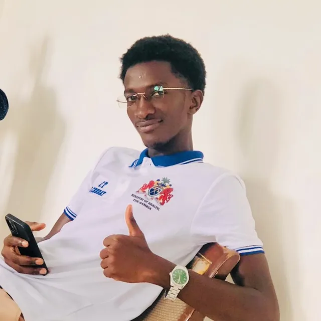 Omar Ceesay in a Ministry of Health polo shirt, leaning back in a chair, thumbs up
